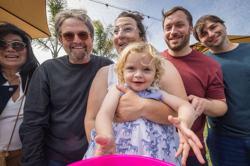 Happy toddler girl held by smiling group of family members at outdoor summer gathering event.