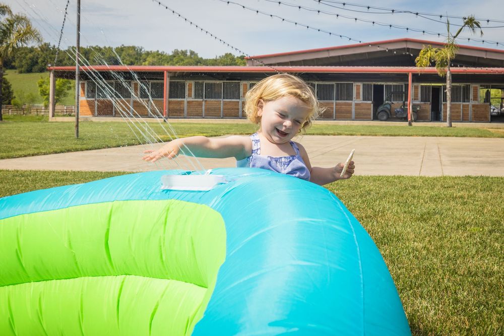 A happy toddler girl in a swimsuit plays on a colorful inflatable water toy on green grass outdoors.