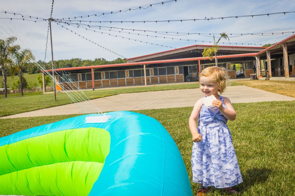Toddler girl in floral dress standing beside colorful inflatable pool on green lawn near rustic building.