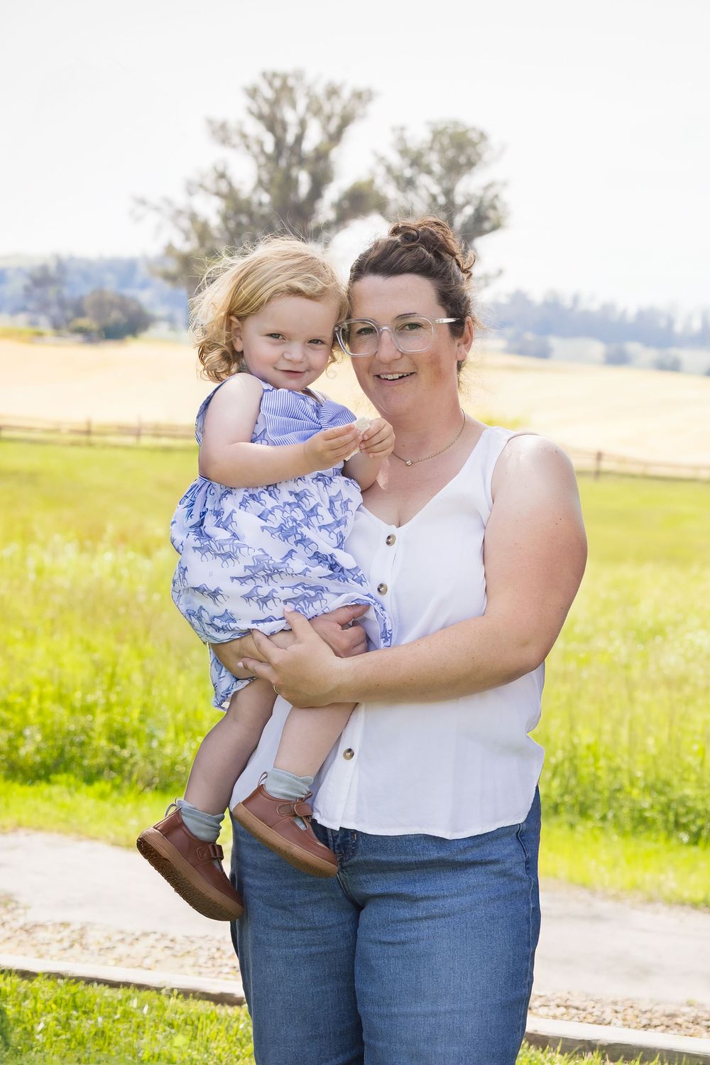 A smiling woman in glasses holds a toddler girl in a blue dress outdoors in a sunny green field.