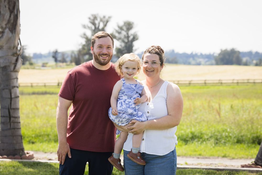 A smiling family of three poses outdoors on a sunny day, with a man, woman, and toddler girl in a blue dress.