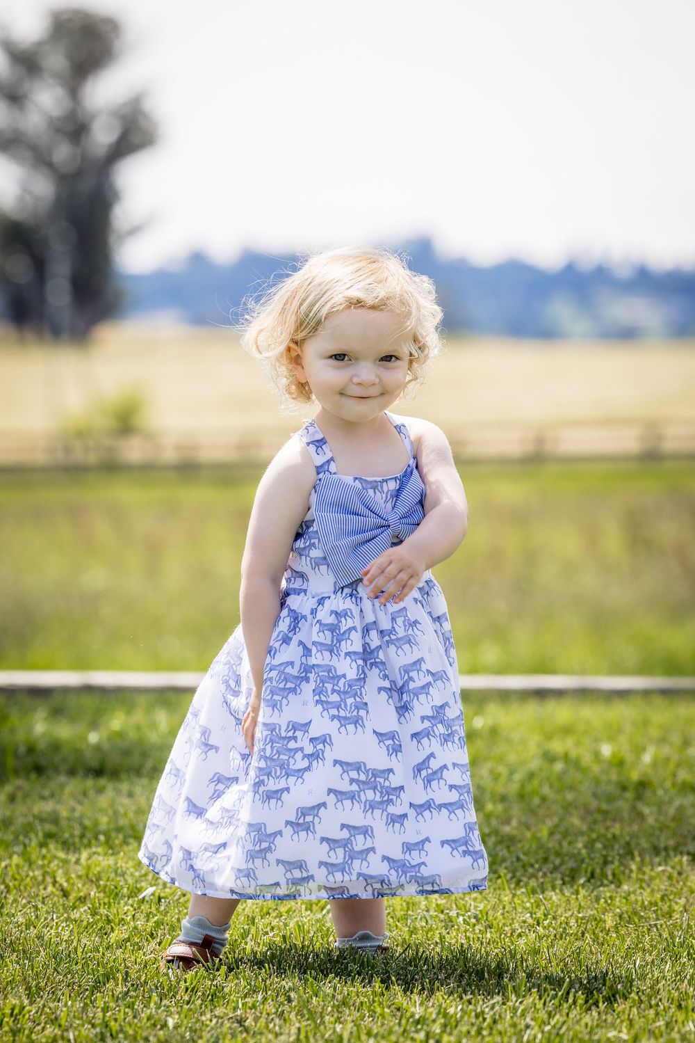 Curly blonde toddler girl smiling in a blue horse-print dress standing on green grass outdoors.