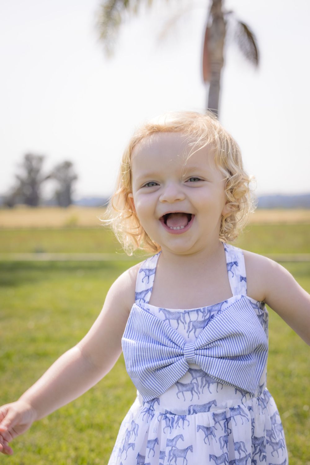 A joyful toddler girl with curly blonde hair laughs outdoors in a blue horse-print dress with a bow.