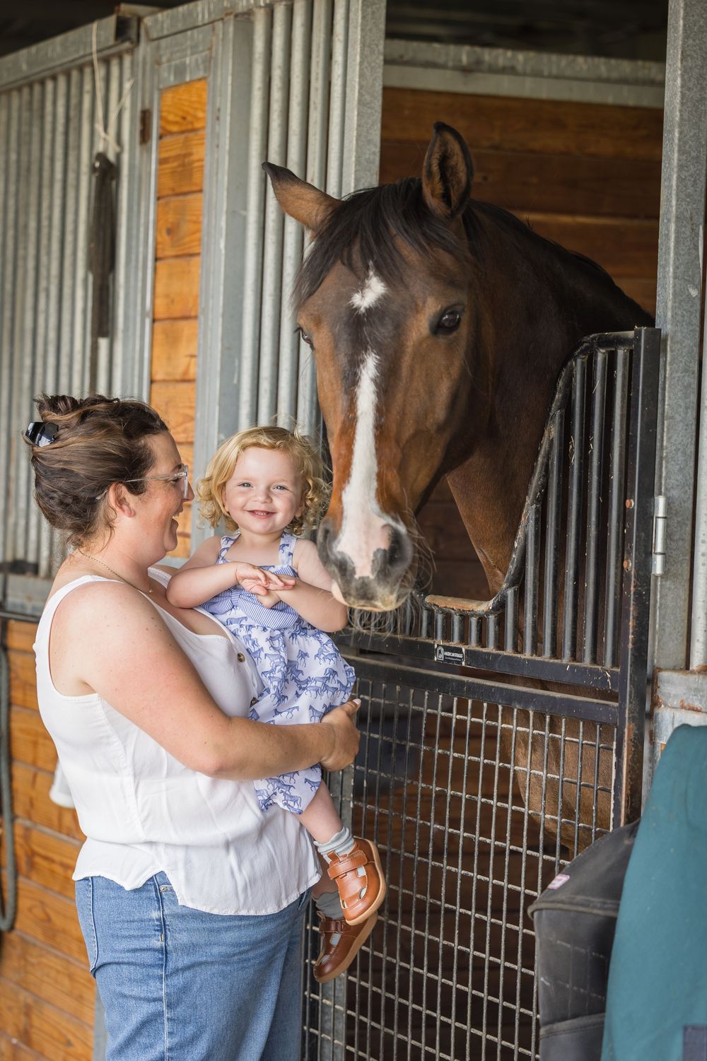 Woman holding smiling toddler girl next to a brown horse in a stable stall.