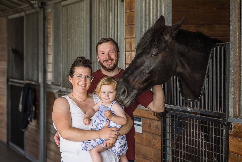 Family with young child posing beside a dark horse in wooden stable stalls.