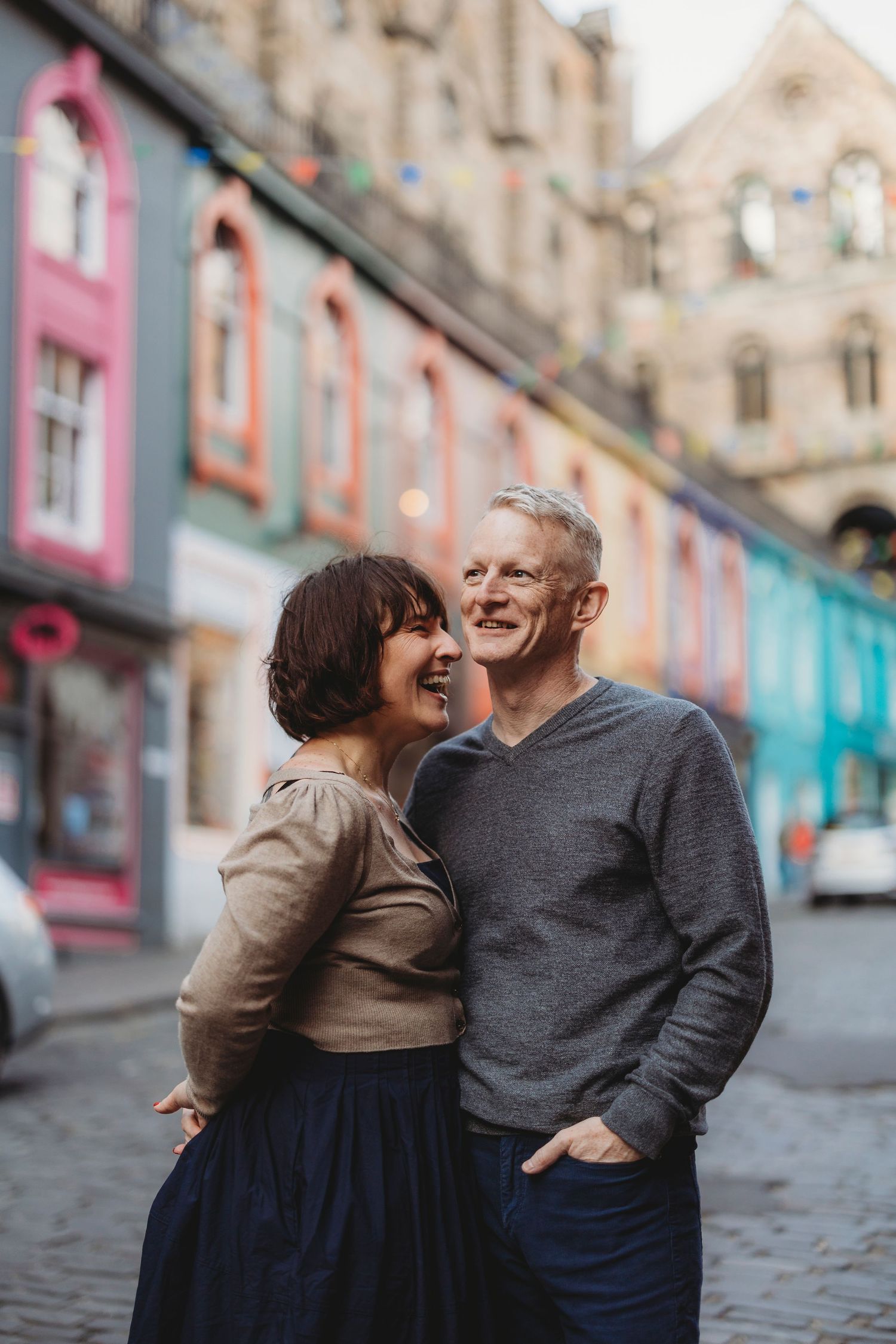 A smiling couple embraces on a colorful cobblestone street with vibrant building facades in the background.