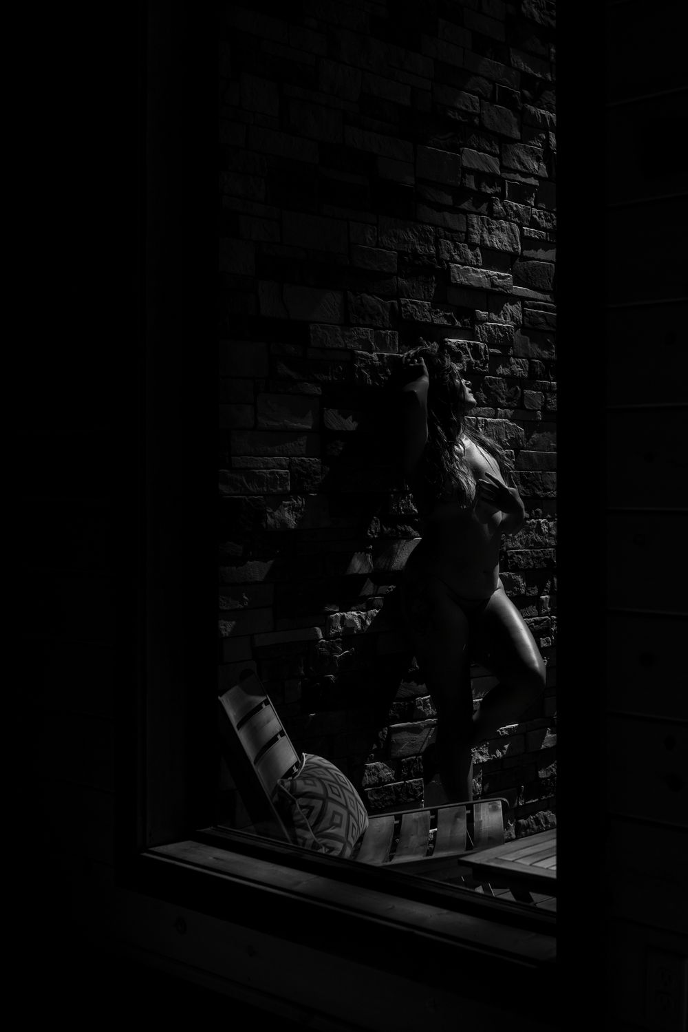 Black and white photo of a figure posed against a brick wall, captured through a dark window frame.