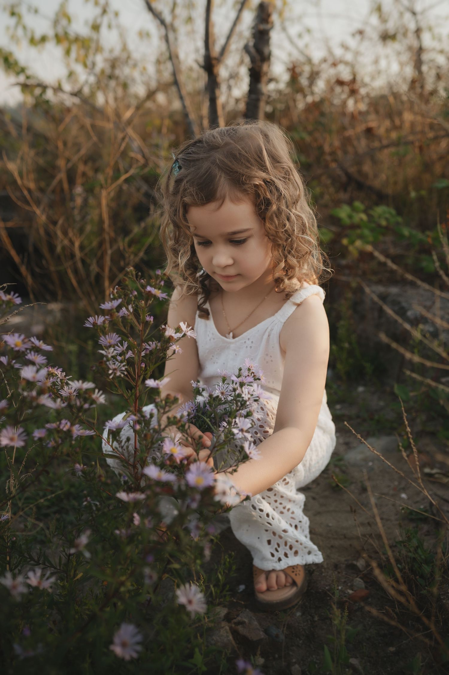 Young girl in white romper crouching to pick purple wildflowers in a natural outdoor setting.