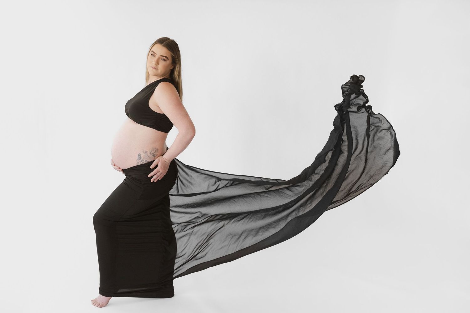 Black and white maternity photo of a pregnant woman in a flowing black dress with fabric billowing dramatically.