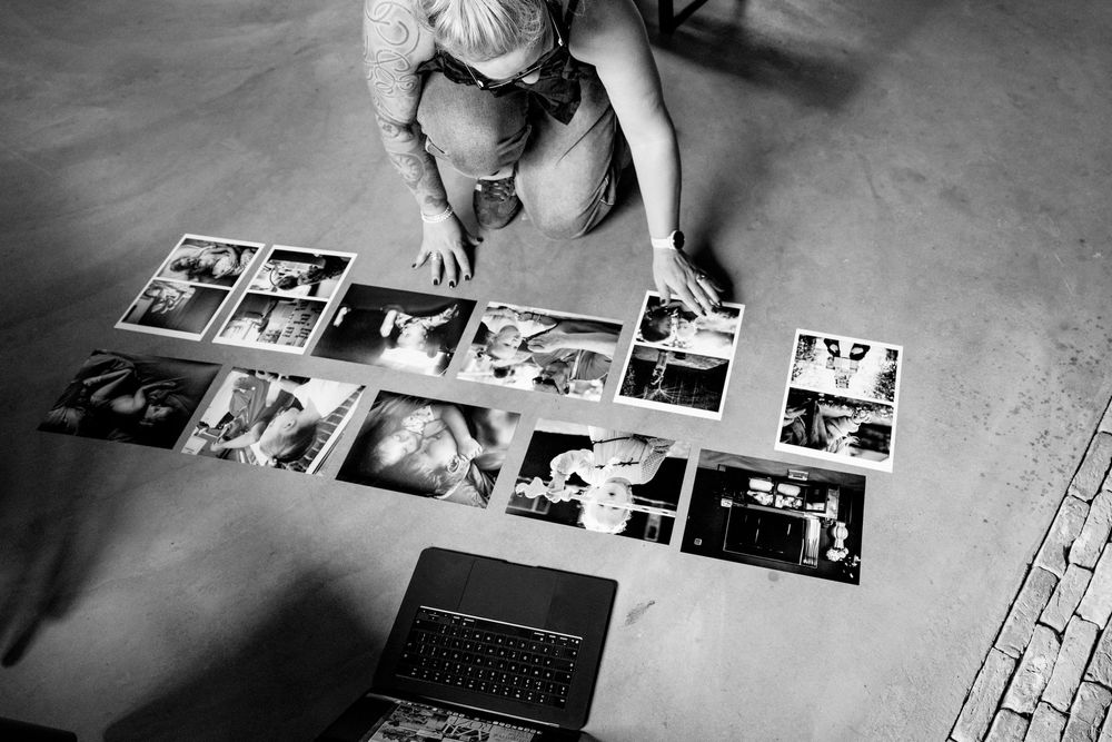 Black and white overhead photo of a person arranging printed photographs on a concrete floor next to a laptop.