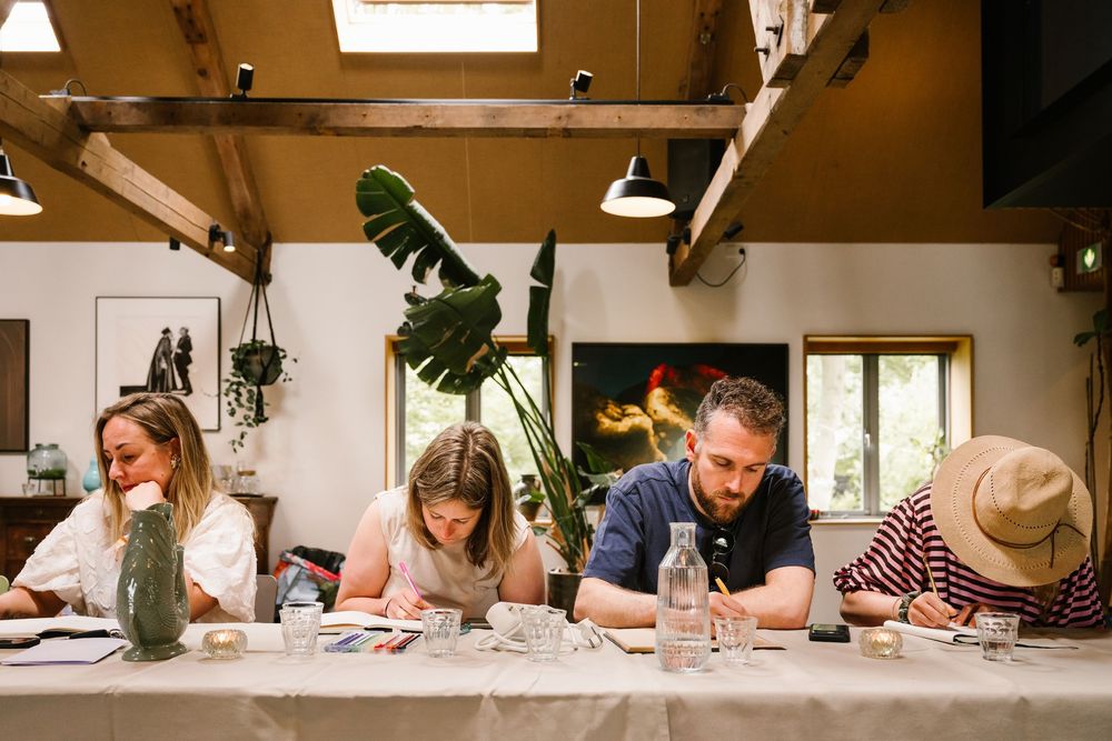 Four people sit at a long table in a rustic studio, focused on drawing or writing during a creative workshop.