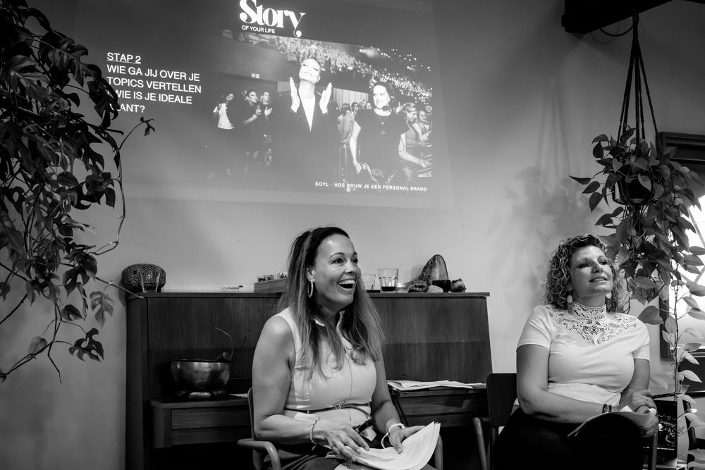 Two women laughing at a Story presentation event, with a projected slide visible in the background.
