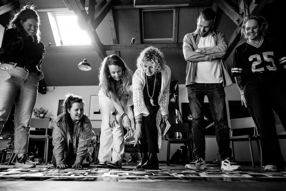 Black and white photo of a group of people gathered around photos spread on the floor in a rustic studio space.