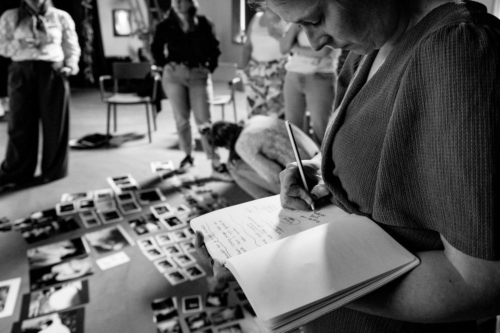 Woman writing notes in journal surrounded by photo prints during creative workshop, black and white.
