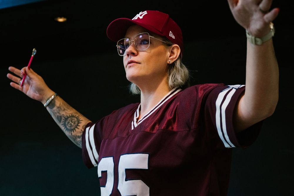 Woman in maroon jersey and cap gestures while holding a pen at an event.