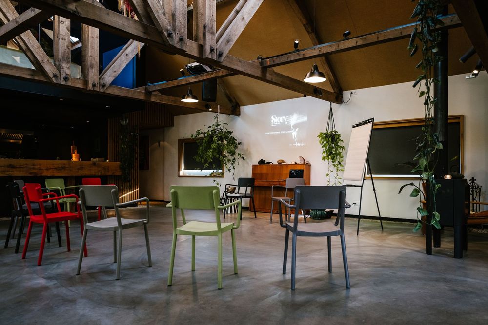 Modern meeting space with colorful chairs arranged in circle, exposed wooden beams, green plants, and whiteboard in industrial loft.