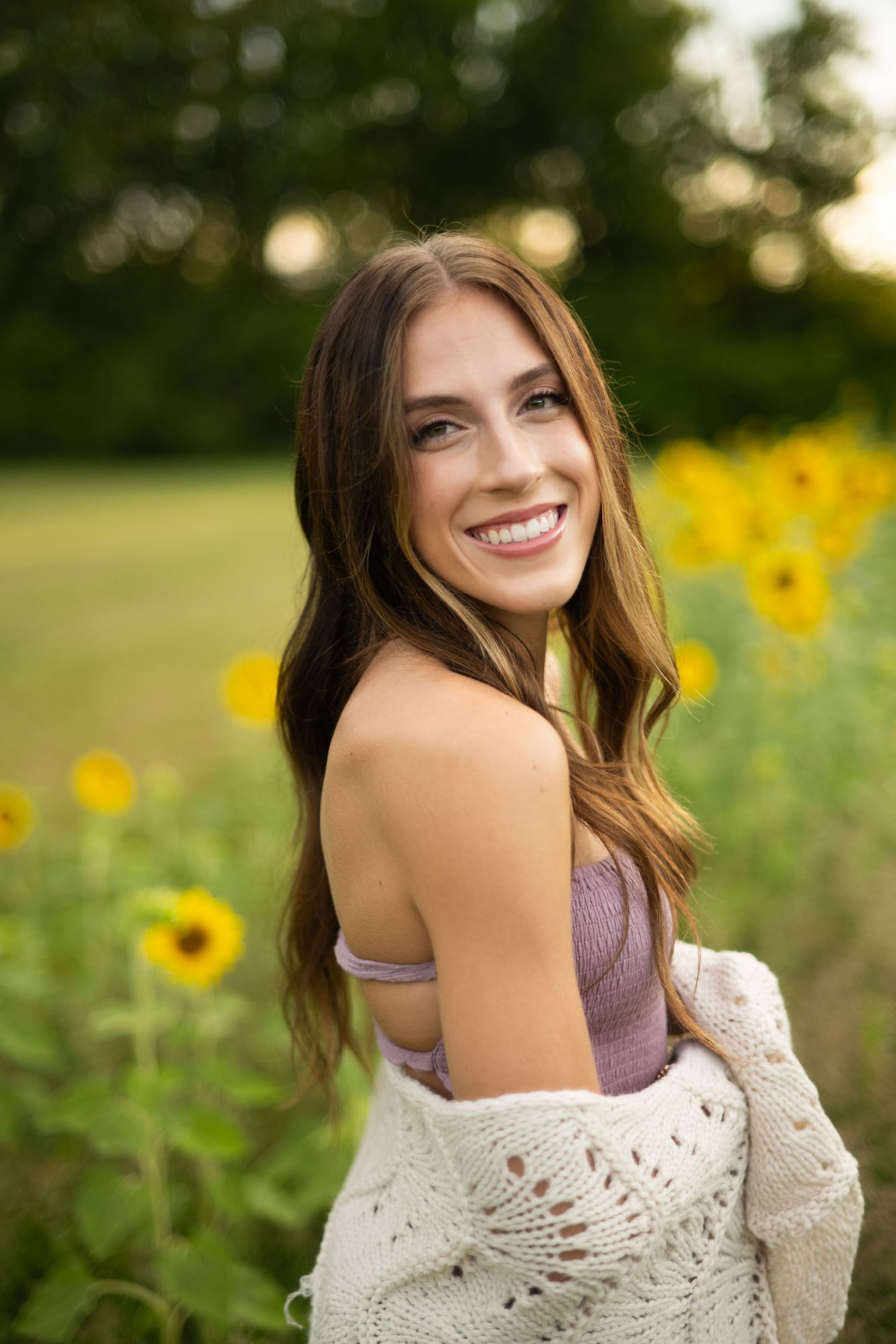 Smiling young woman with long brown hair in a sunflower field, wearing a pink top and white knit cardigan.