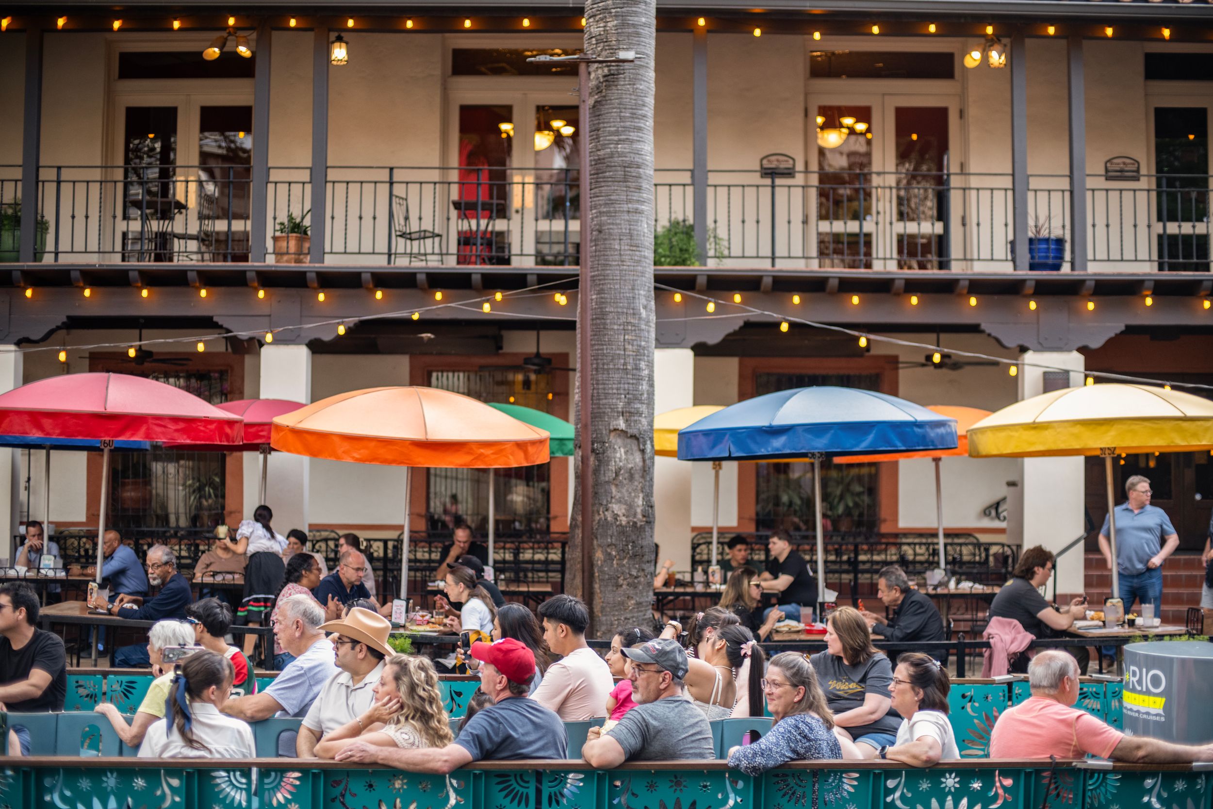Crowded outdoor restaurant patio with colorful umbrellas and string lights on a lively two-story building.