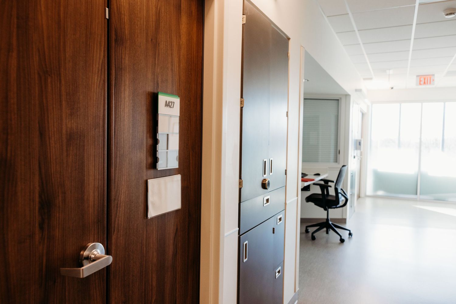 Modern office hallway with wooden door, keypad entry, storage cabinets, and open workspace with chair visible.