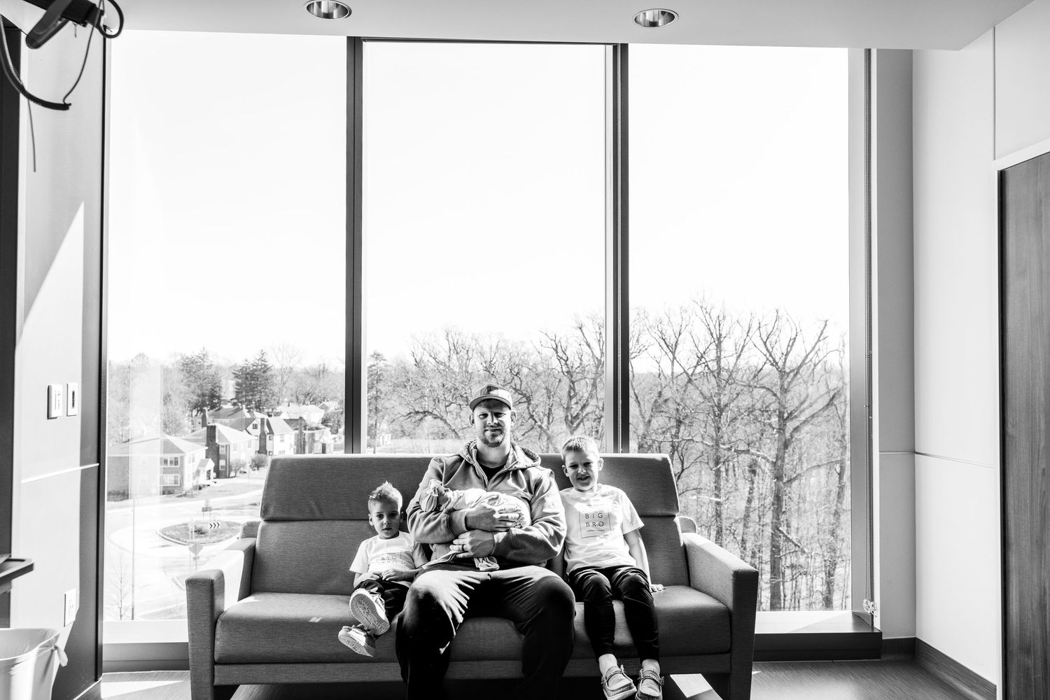 Black and white photo of a family sitting together on a couch by large windows in a bright modern room.