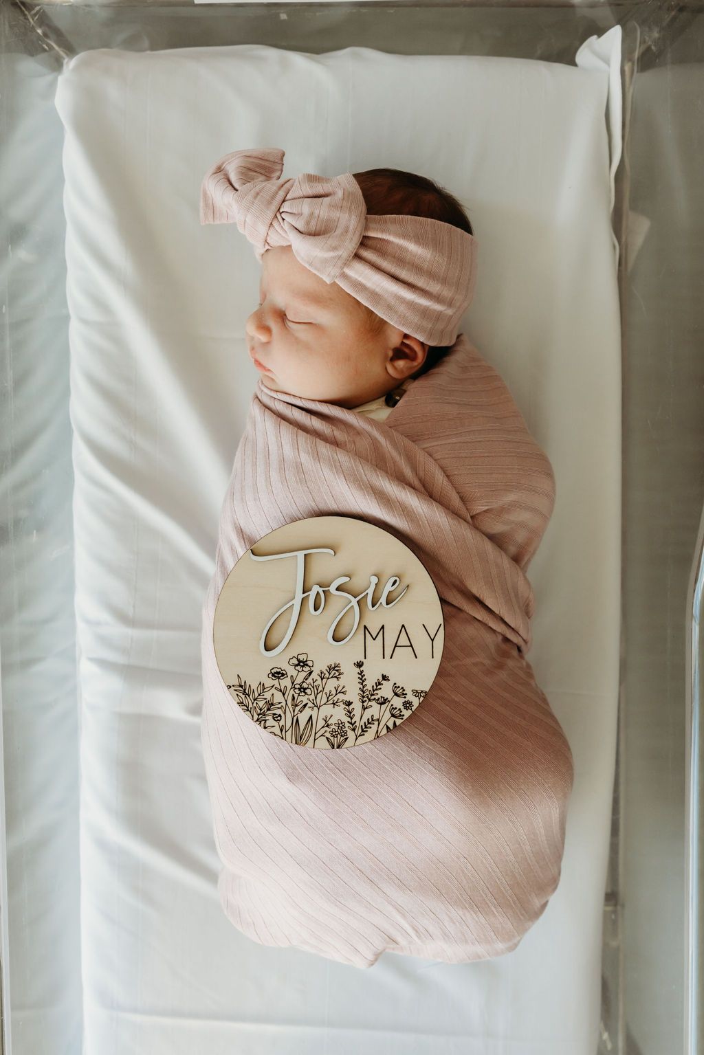 Newborn baby girl swaddled in pink wrap with bow headband, lying in bassinet with wooden name sign reading Josie May.