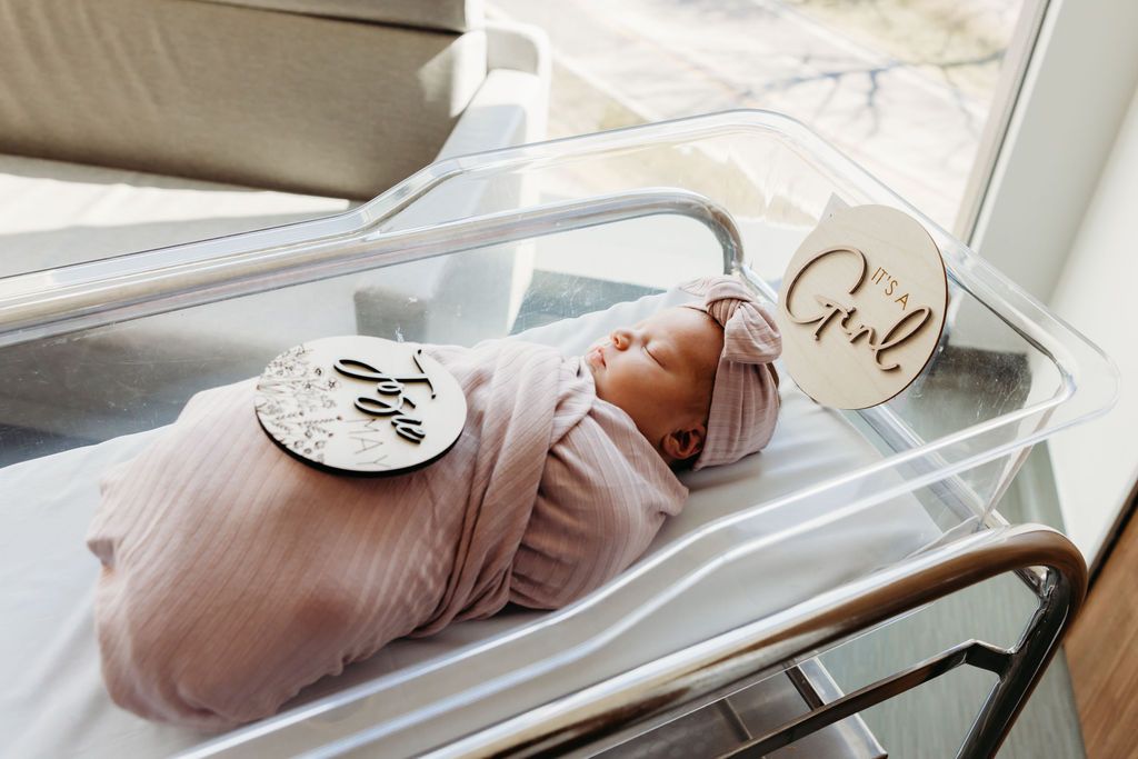 Newborn baby swaddled in pink wrap lying in hospital bassinet with milestone discs.