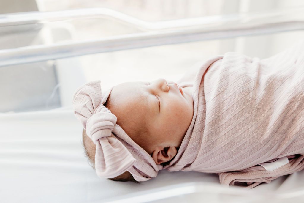 Peaceful newborn baby swaddled in a pink striped blanket with a matching bow headband sleeping in a hospital bassinet.