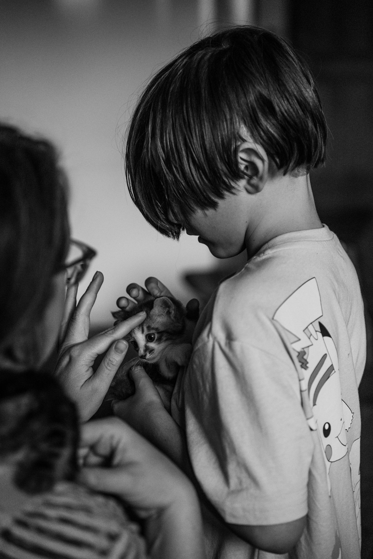 Black and white photo of a young boy in a Pikachu shirt holding a tiny kitten with an adult nearby.