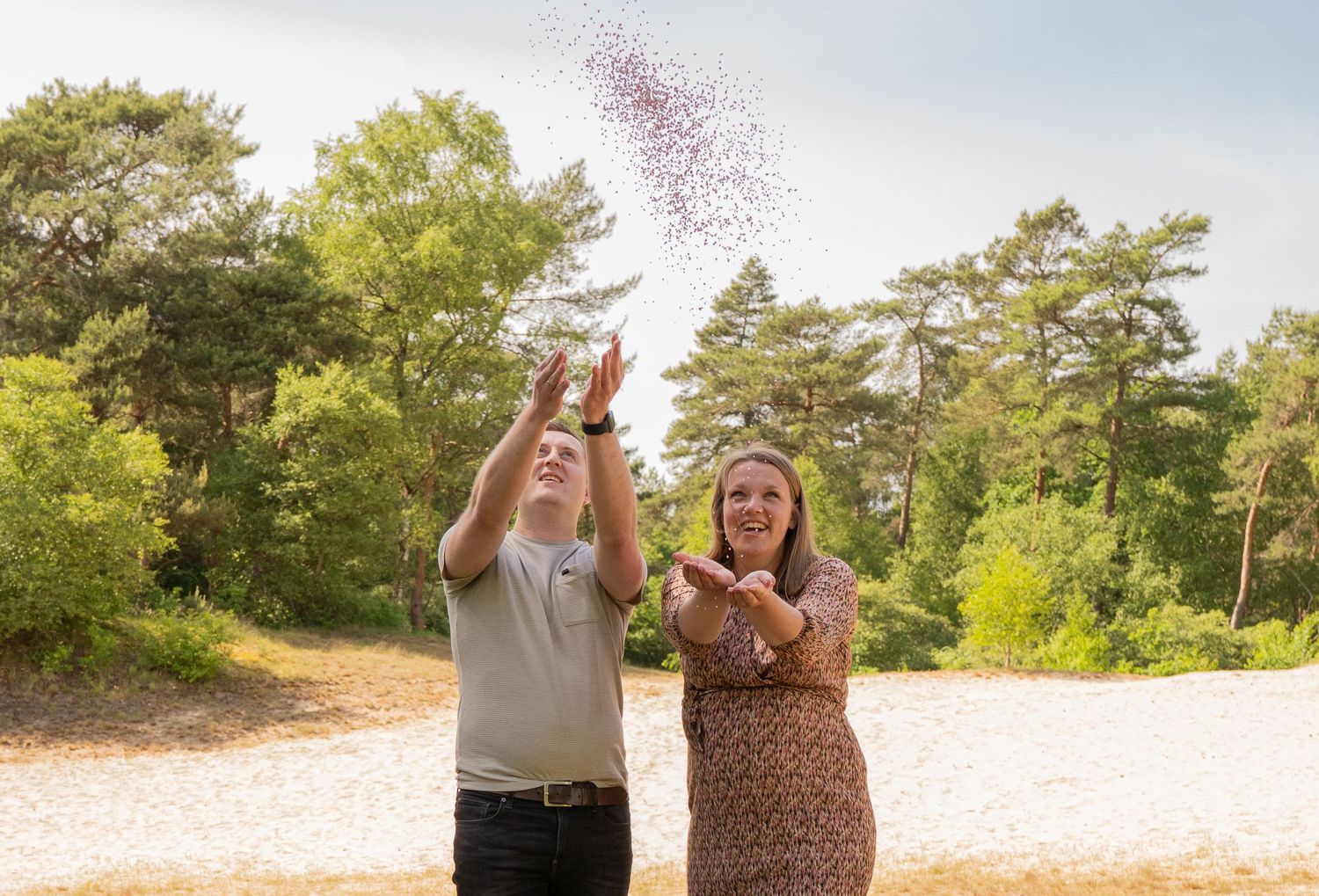 Een stel strooit vrolijk roze confetti in de buitenlucht tussen de dennenbomen op een zonnige dag.