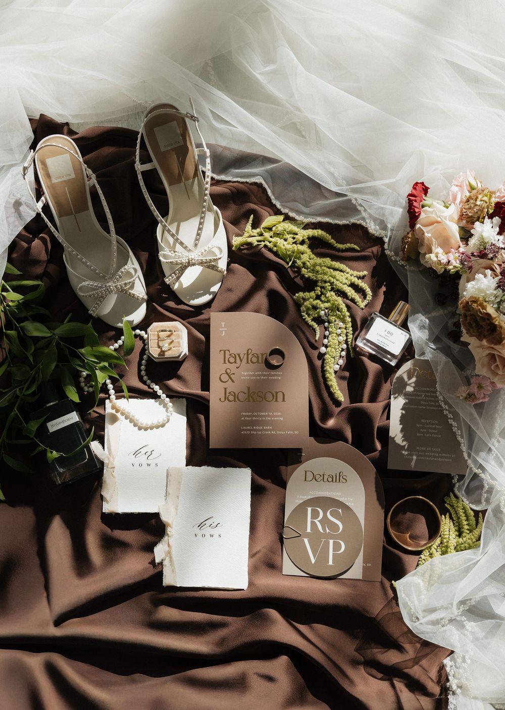 Wedding flat lay with brown invitations, white heels, pearl jewelry, rings, and floral bouquet on silk.