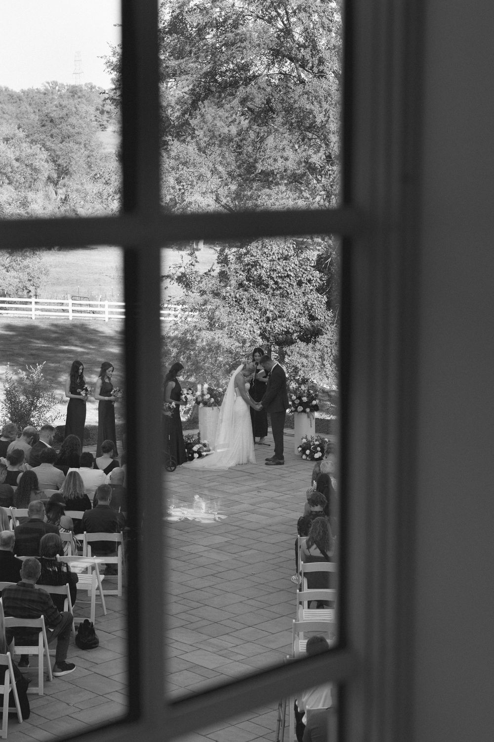 Black and white photo of an outdoor wedding ceremony viewed through a window, with guests seated and couple at the altar.
