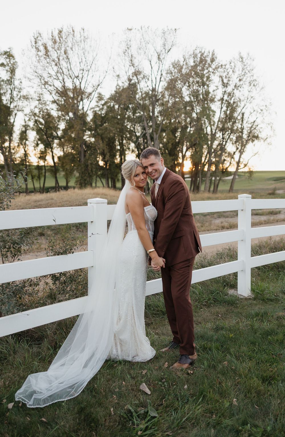 Bride and groom pose by white fence at golden hour, she in lace gown with veil, he in brown suit.
