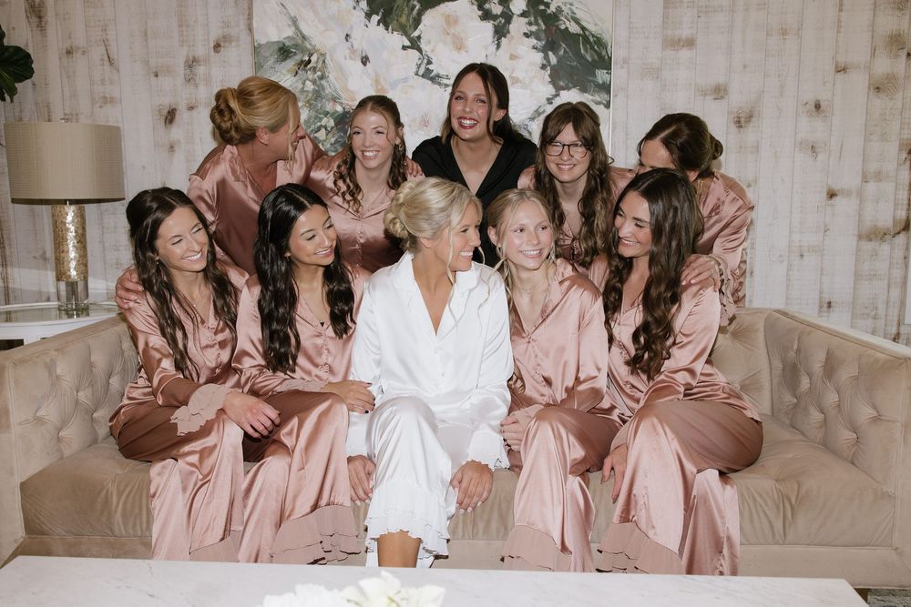 Bride in white robe surrounded by bridesmaids in blush satin robes sitting together on a tufted sofa.
