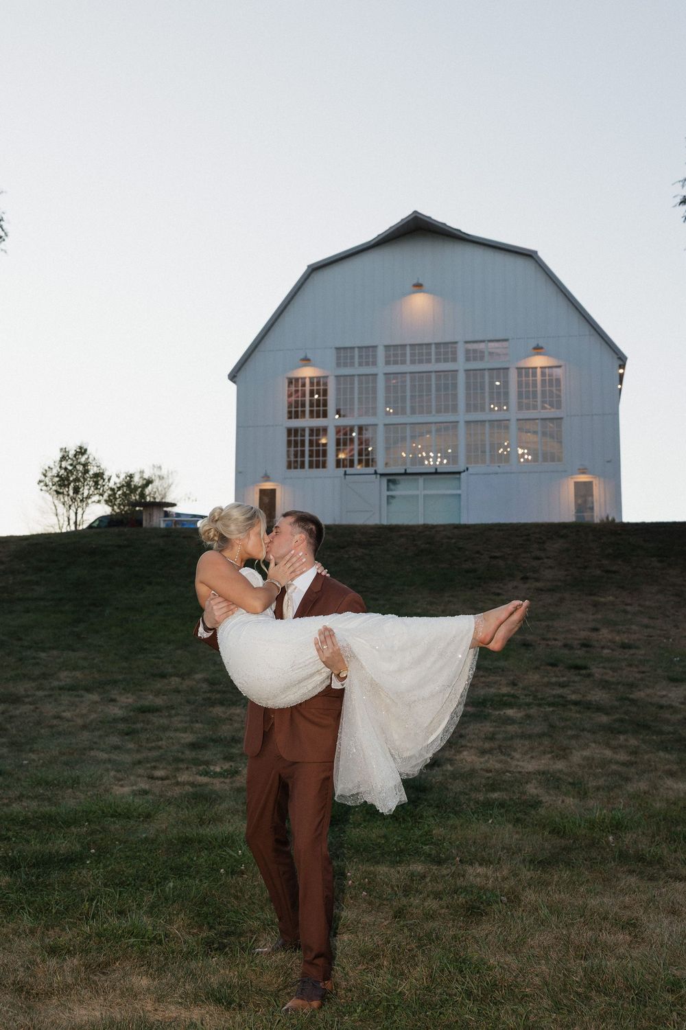 Groom lifts bride in front of a lit white barn venue at dusk in a romantic wedding photo.