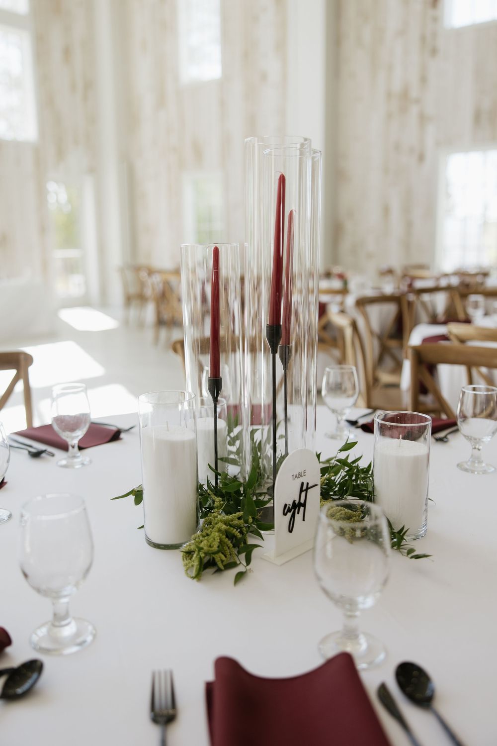 Elegant wedding reception table with tall burgundy taper candles, greenery centerpiece, and burgundy napkins.