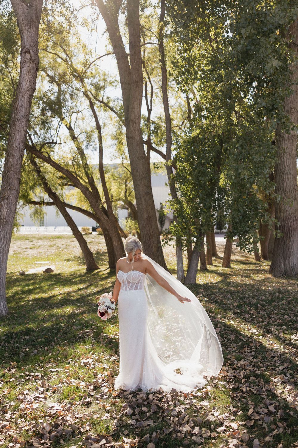 Bride in a flowing white strapless gown and veil walking among autumn trees holding a bouquet.