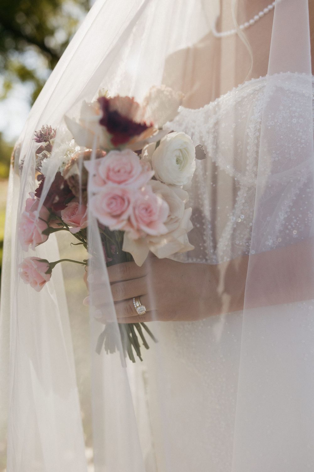 Close-up of bride holding pink and white rose bouquet with engagement ring visible through flowing veil.