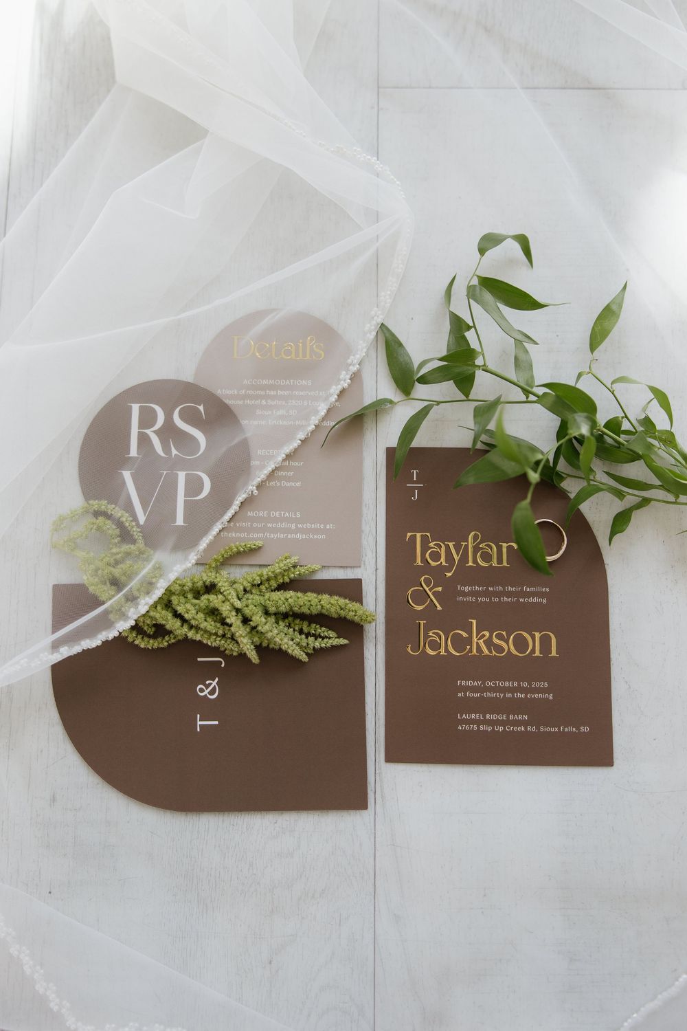 Modern brown and gold wedding invitation suite with RSVP card, greenery, and a ring on white marble.