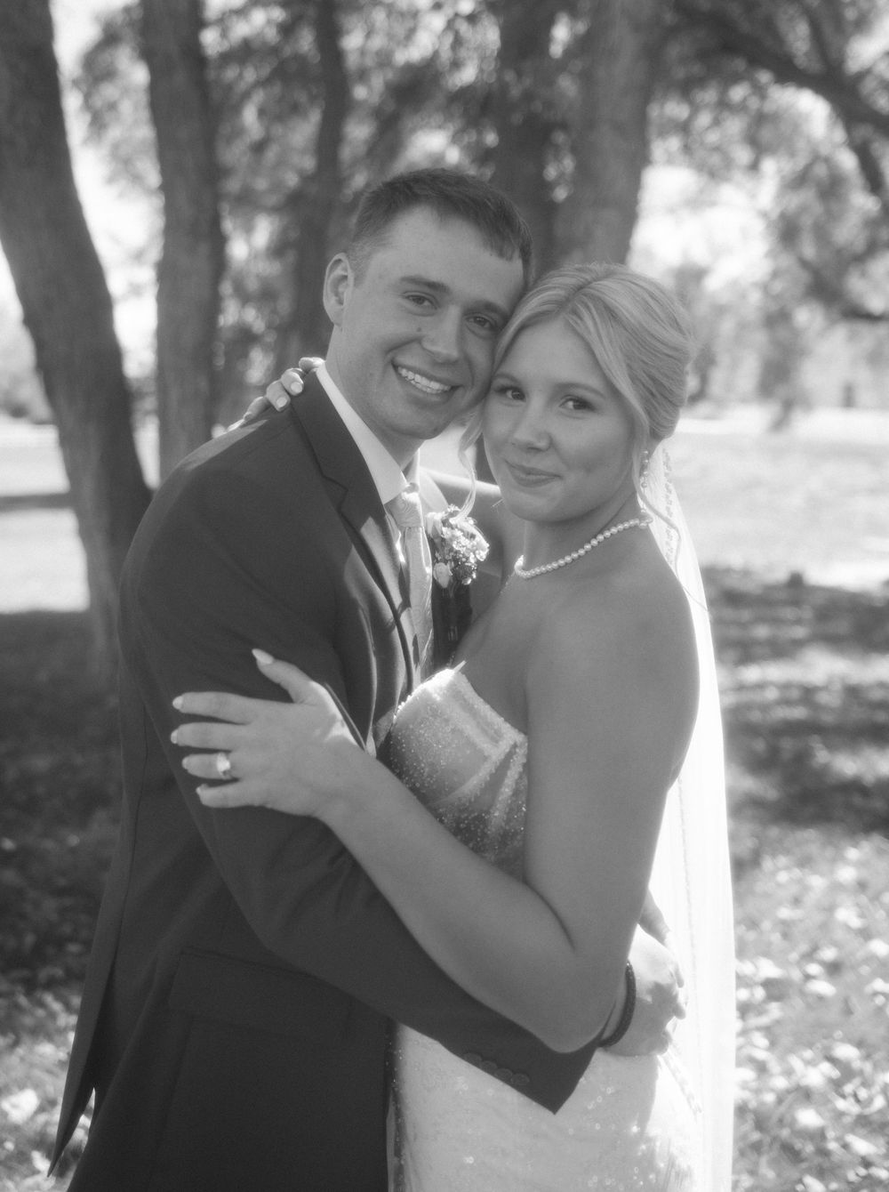 Black and white portrait of smiling bride and groom embracing outdoors under trees on wedding day.