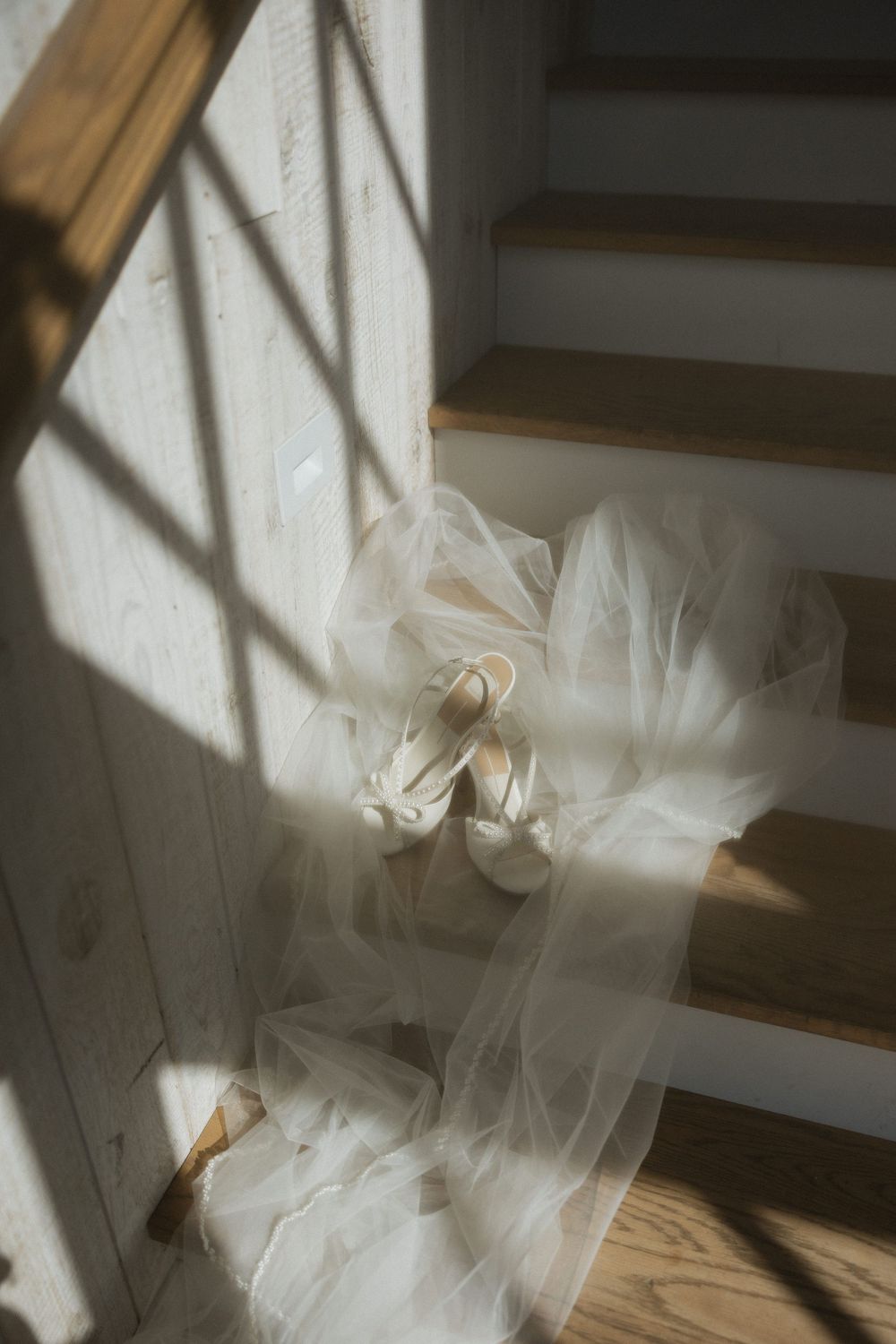 Bridal veil and white heeled shoes arranged on wooden staircase in soft natural window light.