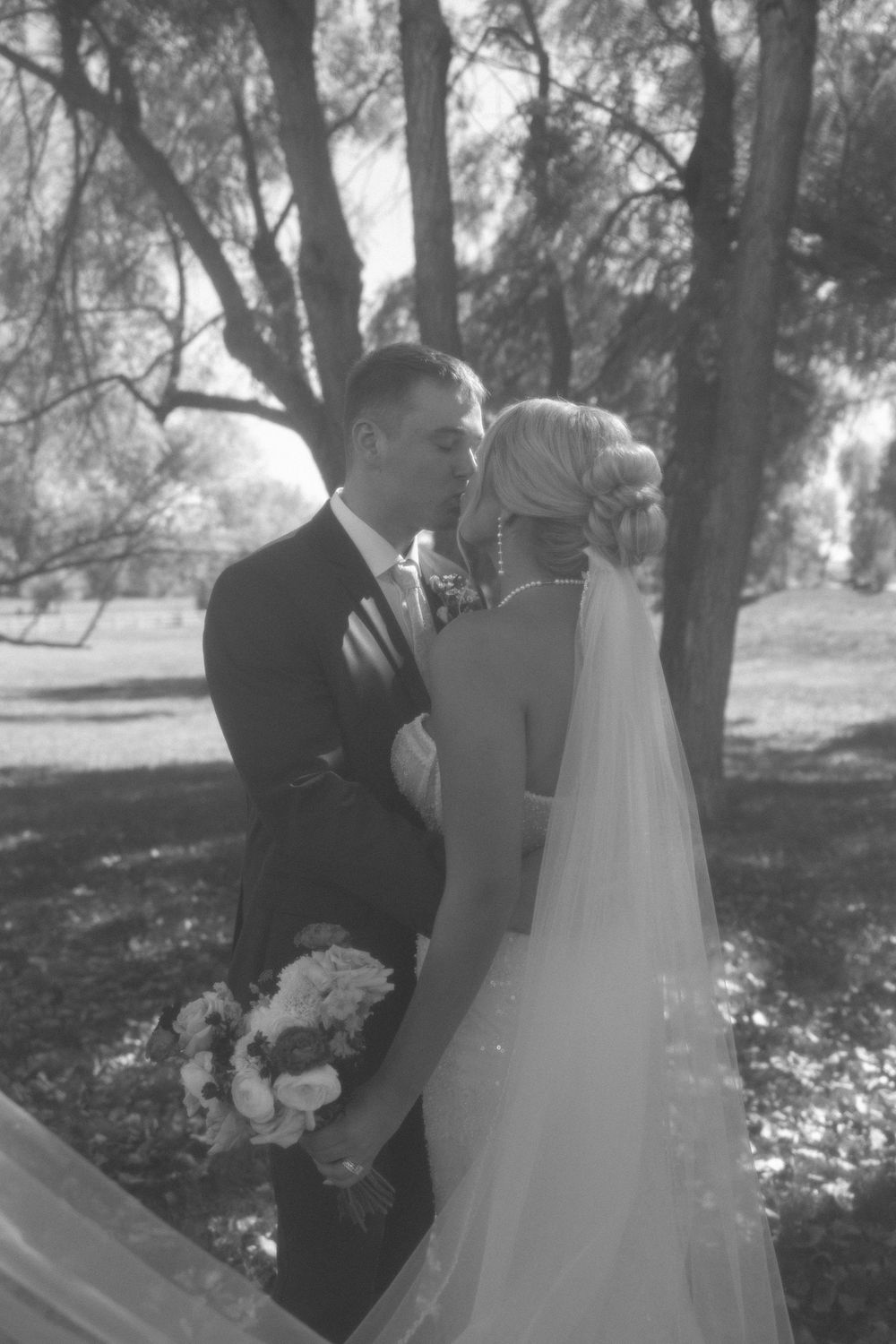 Black and white photo of bride and groom sharing a tender kiss among tall trees outdoors.