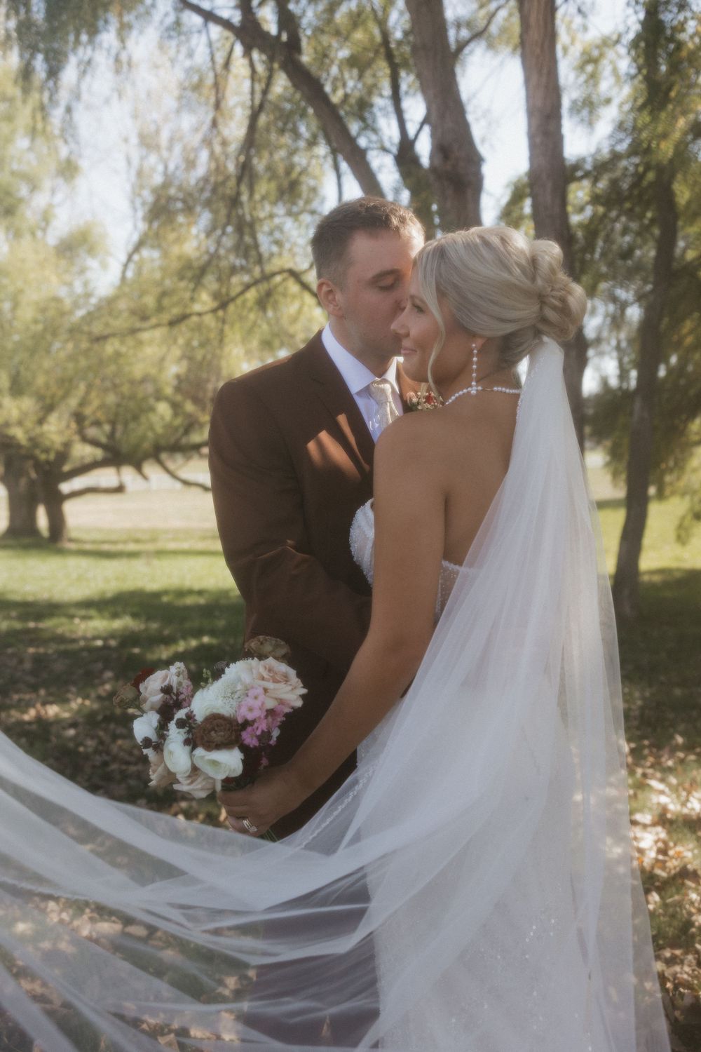 Bride and groom sharing a romantic kiss surrounded by flowing veil and lush trees in golden light.