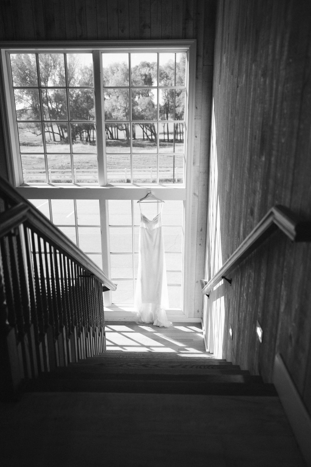 Wedding dress hanging in a stairwell window in black and white, casting soft light on wooden stairs and railings.