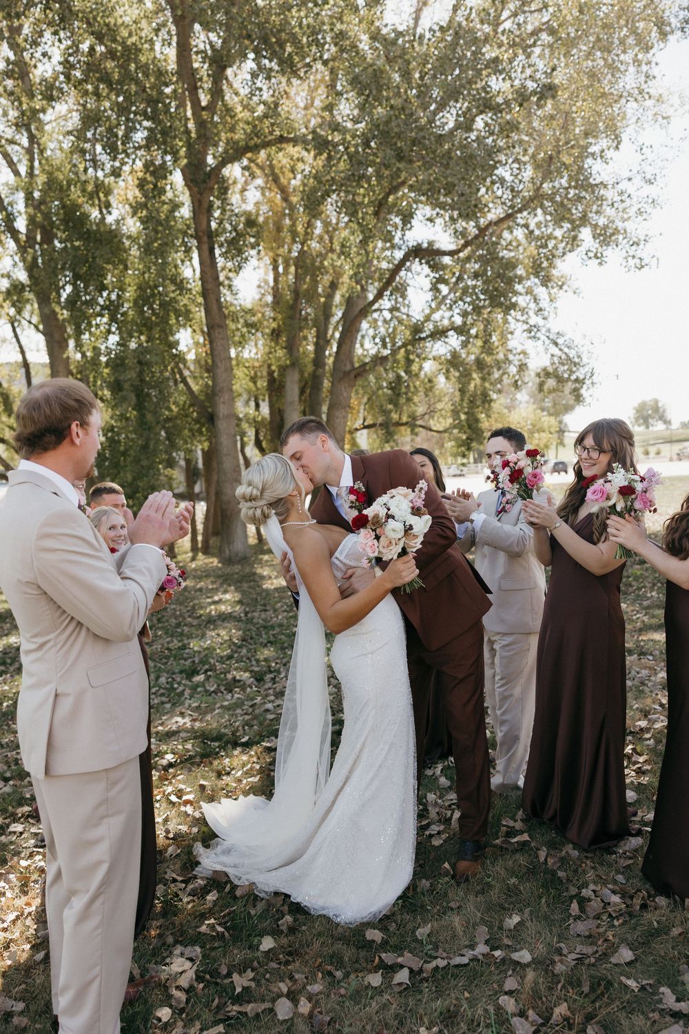 Bride and groom kissing while wedding party cheers and celebrates among autumn trees outdoors.