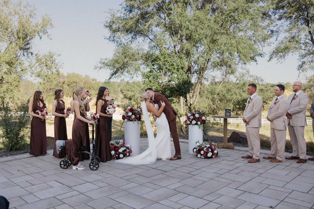 Outdoor wedding ceremony with bridesmaids in burgundy dresses and groomsmen in tan suits as couple shares a kiss.