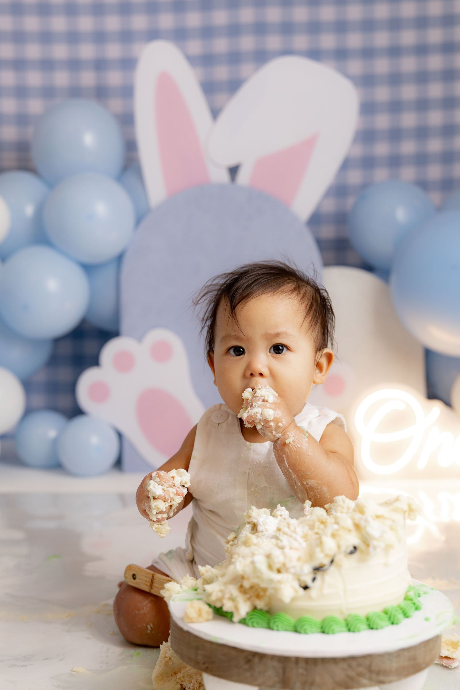 Baby enjoying first birthday cake smash with blue balloons and bunny decorations in the background.