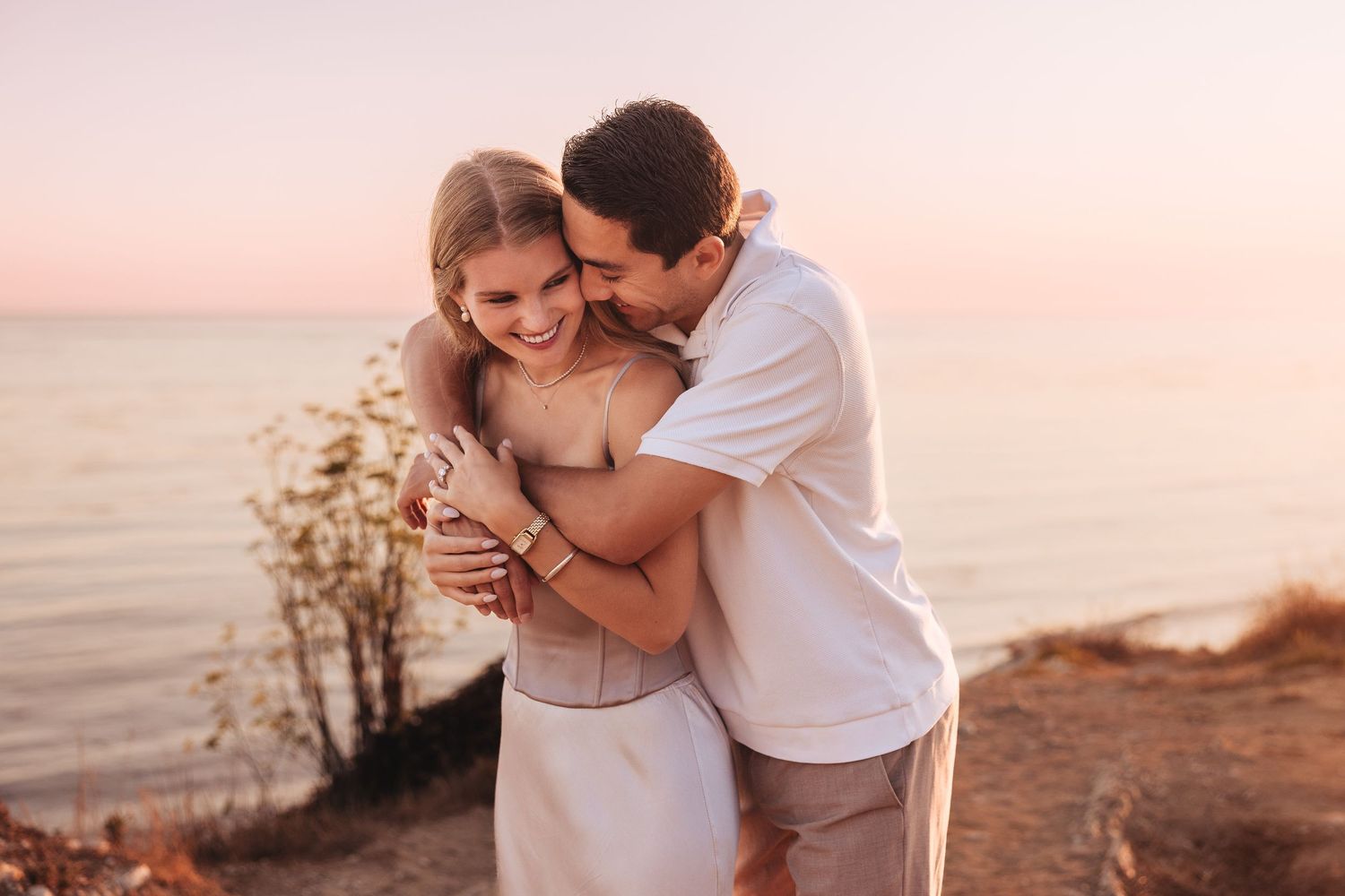 A couple embraces romantically at sunset by the ocean, the woman smiling joyfully in a white dress.