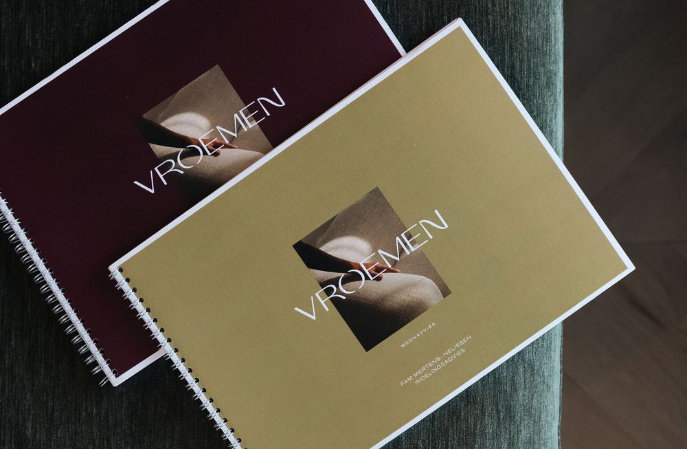 Two spiral-bound VROEMEN branded booklets in gold and burgundy covers resting on a teal surface.