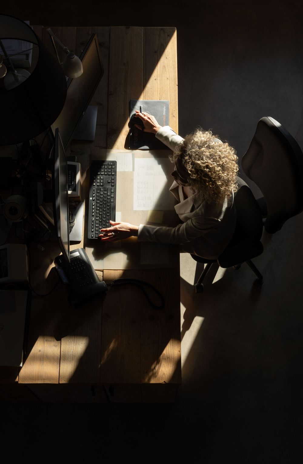 Aerial view of a curly-haired woman working at a wooden desk with a keyboard in dramatic natural light.