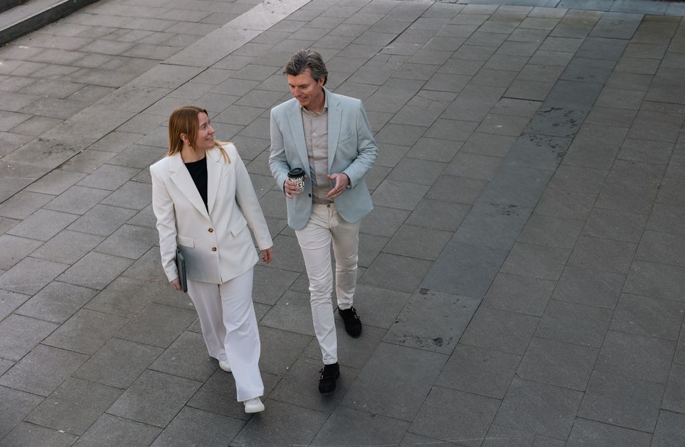 Two business professionals in light suits walking and talking outdoors on a gray paved plaza.